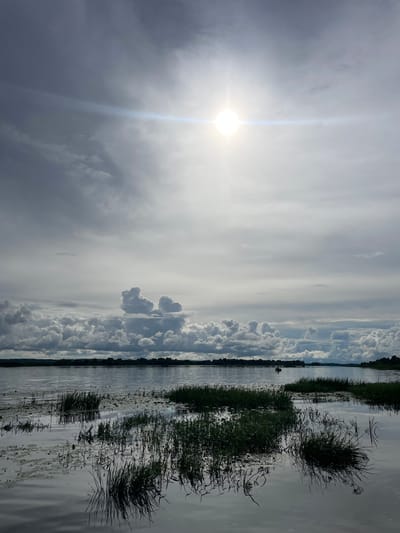 The image depicts a wide, calm body of water, likely a river or lake, with dense vegetation growing along the foreground edges. The water reflects the overcast sky, which is dominated by dramatic, textured clouds. The sun is partially obscured by the clouds but shines brightly, creating a lens flare effect. In the distance, a treeline forms the horizon, and a small boat with at least one person is visible on the water. The overall scene suggests a natural landscape, possibly in or near Makar'evo, Russia, given the provided context. The lighting is subdued due to the cloud cover, but the sun's presence adds a radiant glow to the upper portion of the sky.