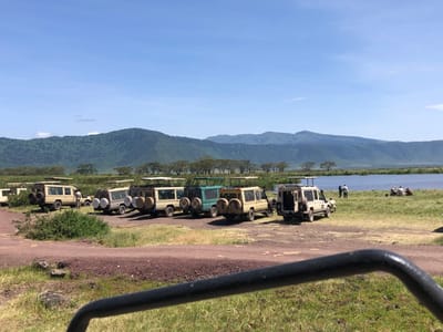 A row of seven safari vehicles are parked on a dirt road, facing a body of water. There are several people standing near the vehicles, and a few are sitting in a grassy area by the water. The scene is set against a backdrop of rolling green hills. The location is the Ngorongoro Crater, a UNESCO World Heritage Site near Ilturot Lorkereyan, Tanzania.