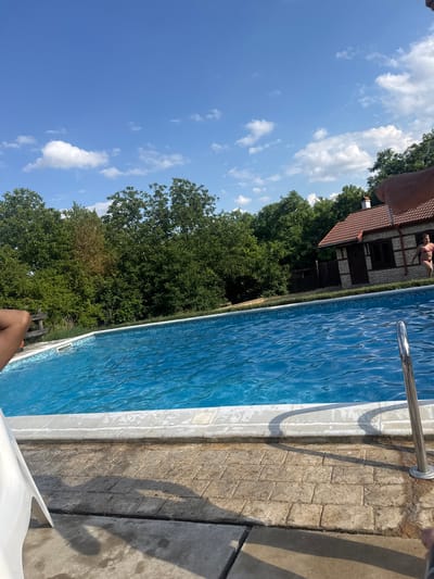 A wide shot captures a swimming pool on a sunny day. The pool, filled with blue water, has a tiled border and is surrounded by a stone patio. A white plastic lounge chair is partially visible in the foreground. In the background, a small stone building with a red tile roof stands amongst lush green trees. A woman in a pink bikini is visible near the building, standing by the pool edge. The sky above is blue with scattered white clouds. The location is identified as Varbak, Bulgaria.
