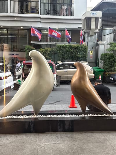 This image captures a street scene in Khan Boeng Keng Kang, Cambodia. In the foreground, two large, stylized bird sculptures are positioned side-by-side on a raised platform. The sculpture on the left is a pale, creamy color, while the one on the right is a metallic bronze. Behind the sculptures, the street is visible with various vehicles, including cars, a tuk-tuk, and possibly other modes of transport. A person in a black shirt is seated with their back to the camera, looking down, located near the bronze sculpture. An orange traffic cone is placed on the ground in front of the bronze sculpture. In the background, a multi-story building with balconies and visible laundry is present. Flags, including what appear to be Cambodian flags, are mounted on the building's facade. A large electrical transformer unit is also visible on the side of the building, bearing the "SEE" logo. Green trash bins are situated on the right side of the image. The overall environment suggests an urban setting.