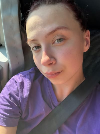 A woman is sitting in a car wearing a purple shirt. She is wearing a seatbelt. The location is unknown.