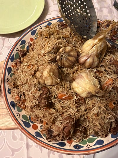 The image provides an overhead view of a large, round serving platter laden with a rich Plov, a traditional rice dish, set on a table. The Plov consists of long-grain rice mixed with pieces of meat, likely lamb or beef, and shredded orange carrots. Several whole, roasted garlic bulbs are prominently placed within the dish, appearing soft and caramelized.
A metal slotted spoon or skimmer is partially visible, resting within the Plov on the left side of the platter, suggesting it's ready for serving. The platter itself has a decorative rim featuring a colorful pattern of blue, orange, and green teardrop-like shapes and lines.
In the bottom left, a section of a plain, light green plate is visible. The table is covered with a white tablecloth that has a subtle, light-colored, ornate patterned design. A wooden surface is also partially visible beneath the tablecloth on the right.
The scene depicts an indoor setting, likely a dining area, where a meal is being served. The lighting appears to be artificial and warm, suggesting an evening meal. The dish's characteristics are consistent with Central Asian cuisine, which is common in Russia, aligning with the provided location context of Пролетарский, Russia. There are no people visible and no visible text in the image.