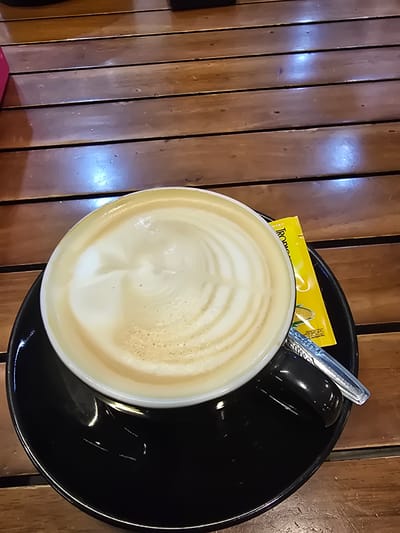 A close-up, high-angle shot shows a cup of latte with foam art resting on a black saucer, which is placed on a wooden table. A yellow packet, likely containing sugar, is visible next to the cup, and a silver spoon rests on the saucer. The wooden table has a visible grain and is composed of several planks. The lighting suggests an indoor setting, possibly a cafe. No people are visible in the image. The location is identified as Ramunia Dua, Indonesia.