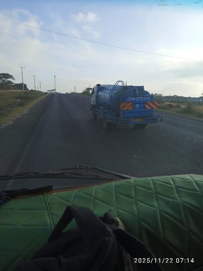 A road stretches into the distance under a partially cloudy sky. On the road, a blue water truck with the number 0794 272 542 and license plate KAU 245U travels in the same direction as the viewer. The view is from inside another vehicle, capturing the dashboard and a green quilted seat cover. The timestamp indicates 2025/11/22 07:14. Utility poles line the road's edge, and sparse vegetation covers the surrounding landscape.