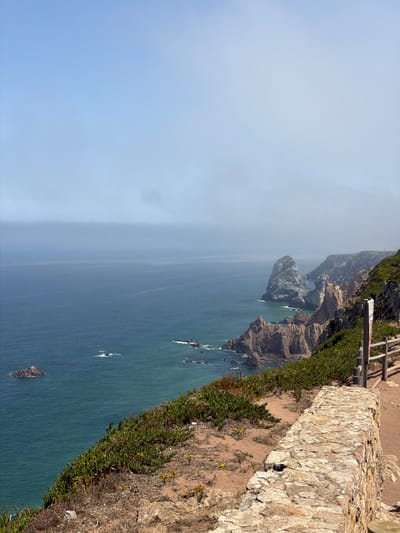 This is a scenic view of the Atlantic Ocean and the dramatic coastline at Cabo da Roca, located near Colares, Portugal. The image captures a steep cliff overlooking the sea, with rugged rock formations jutting out from the water. The ocean is a deep blue-green, with waves gently crashing against the rocks. The cliff face is covered in lush green vegetation, with some patches of dry earth and small yellow wildflowers. A stone wall and a wooden fence line the edge of the cliff, indicating a pathway or viewing area for visitors. The sky is a pale blue with a thin layer of mist or fog hanging low over the horizon, softening the distant views of the sea and sky. There are no people visible in the image. The scene is characterized by natural beauty and a sense of rugged, windswept terrain.