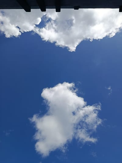 The image displays a vibrant, deep blue sky with scattered white cumulus clouds. At the top of the frame, a dark, shadowy structure, possibly a building overhang or roofline, partially obscures the sky. The clouds are predominantly white, with some exhibiting areas of slight grayness, suggesting varying densities and light penetration. One large cloud formation is centrally located in the lower half of the image, appearing voluminous and textured. Another, larger cloud formation stretches across the upper portion of the sky, interacting with the dark structure above. The overall impression is of a bright, clear day with a few puffy clouds. The location is identified as Lebbae, Indonesia. There are no people or discernible actions depicted in the image.