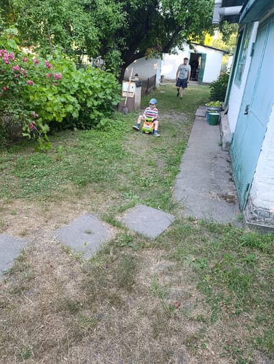 In the city of Ozera, Ukraine, a yard scene unfolds under a canopy of green trees. In the foreground, a young boy wearing a striped shirt and blue and white cap sits on a yellow and orange toy car. To his right, a paved path leads towards a white building with a light blue door. Further back, a young man in a gray t-shirt and plaid shorts walks away from the camera. The yard is covered with grass, some of which appears dry and brown, interspersed with patches of green clover. A bush with pink flowers is visible on the left side of the image, and a large tree with green leaves dominates the upper portion. Several concrete slabs are laid on the ground. The overall impression is of a casual outdoor setting in a rural or semi-rural area.