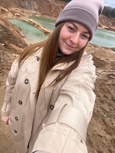 A selfie of a woman with light skin, wearing a grey beanie and a beige trench coat, outdoors. She is smiling at the camera. The background reveals a landscape featuring multiple small turquoise-colored lakes or ponds. The surrounding terrain appears to be a rugged, earthy environment, possibly a quarry or similar industrial site. The location is likely Ivlevo, Russia.