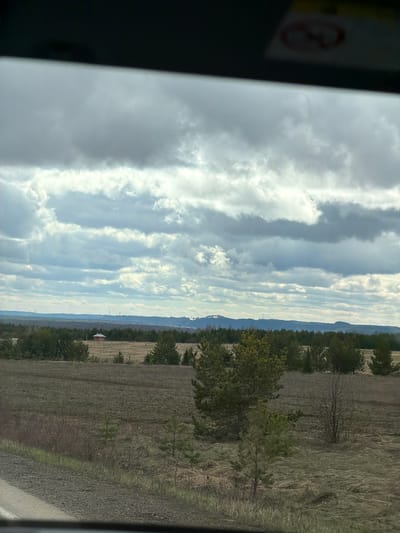 This media file presents a vertical landscape orientation, depicting a rural or natural setting under a cloudy sky. The overall impression is of a quiet, expansive daytime scene.
**Subjects:** The primary subjects include a vast sky, a distant mountain range, rows of evergreen trees, and large, dry fields. In the upper left portion, a blurred section of what appears to be a vehicle's interior or window frame is visible.
**Activity or event taking place:** No specific human or animal activity is depicted. The image captures a static, serene view of the landscape, possibly observed from a moving vehicle.
**Notable details:** The sky dominates the upper two-thirds of the image, filled with dramatic, long, horizontal bands of clouds that create a layered effect. Patches of brighter blue sky are visible between the cloud formations. The lighting suggests a daytime shot, with diffused sunlight. On the distant horizon, a long, hazy blue line of mountains or hills stretches across the frame. Faint, slender structures, possibly distant communication towers, can be seen against this backdrop. A line of dense evergreen trees forms a visual barrier across the midground. Below these trees and extending into the foreground are extensive fields, characterized by dry, brown, and yellowish vegetation, suggesting either fallow land or early spring conditions. A small building with a distinctive red roof is faintly visible in the far left of the field area. The blurred dark area on the left edge indicates the photo was likely taken from inside a structure or vehicle, looking out.
**Visible text:** Within the blurred, dark area in the upper left corner, a circular sign is partially visible. It features a red border and a diagonal red line across its center, consistent with a universal "no entry" or "forbidden" symbol. No specific text is legible on this sign.
**Location:** The scene is located in Povarenki, Russia.