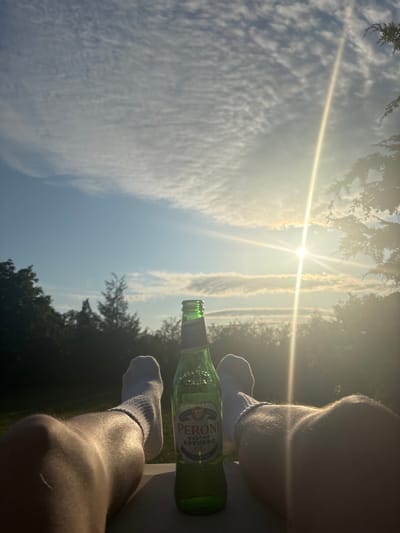 A person wearing white socks relaxes outside on a sunny day, legs extended. A green Peroni beer bottle rests between their feet. The sky is partly cloudy with a bright sun positioned in the upper center of the frame. Green trees are visible in the background, adding to the outdoor ambiance. The image suggests a leisurely, relaxing moment in nature.