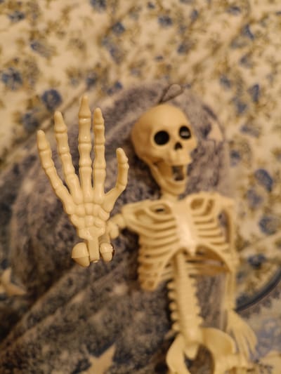 The image shows a plastic skeleton model lying on a blue and white patterned fabric. The skeleton is light beige and appears to be a prop, possibly for Halloween or a decorative item. Its right hand is raised, with fingers spread apart in a gesture that could be interpreted as a wave or a stop sign. The skeleton's mouth is open, giving it a somewhat startled or surprised expression. The background fabric has a floral or abstract pattern with blue and olive green elements on a cream-colored base. The lighting suggests it is daytime, but no specific time can be determined. There is no visible text in the image. The setting appears to be indoors, with the fabric possibly being a blanket, tablecloth, or upholstery. The image is slightly out of focus, particularly in the background, drawing attention to the skeleton. The overall tone is playful or slightly spooky, given the subject matter.