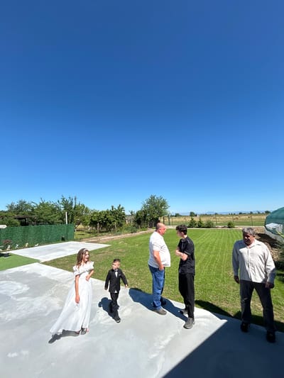 A sunny outdoor scene, possibly a wedding or formal event, shows a group of people on a white concrete or tiled area. A woman in a white dress stands near a boy in a suit. Further away, a man in a white shirt and jeans stands with his arms folded. Next to him is a person in a black shirt and pants. An older man in a light-colored shirt and dark pants is at the far end of the concrete area. They are all standing on a well-maintained lawn with a fence and trees behind them. The sky is clear blue. The location appears to be in Okop, Bulgaria, based on the presence of typical Bulgarian flora and fauna.
