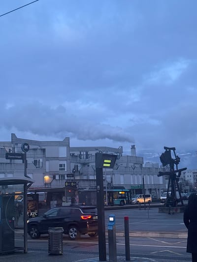 A cityscape under a cloudy sky. A street with traffic, including a bus and car. A KFC establishment is visible. A metro station named "Étienne Grappe" is also present. A unique sculpture stands amidst the buildings. The sky is overcast with hues of gray and blue.