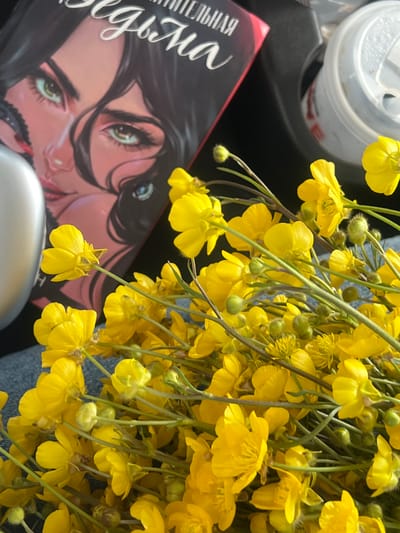 The image shows a book with a portrait of a woman on the cover, a half-full beverage can, and a bouquet of yellow flowers, possibly buttercups. The overall setting appears to be indoors, likely a table or lap. No details indicate the location to be Apnagyugh, Armenia.