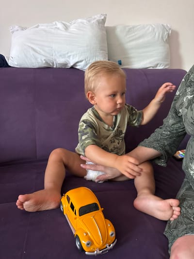 The image shows a young child lying on a purple surface, possibly a couch or bed, with several white pillows beside them. The child is wearing a camouflage-patterned t-shirt and a diaper. Their right arm is raised, and their left hand is resting on their leg. A bright yellow toy car is positioned next to the child's feet. In the background, a person's legs, clad in green camouflage pants, are visible, suggesting the child is with an adult. The scene appears to be indoors, with soft lighting indicating it might be daytime. The overall mood seems relaxed and casual, with the child looking towards the right side of the frame. There is no visible text in the image. The location context of Sochi, Russia, suggests a domestic setting.