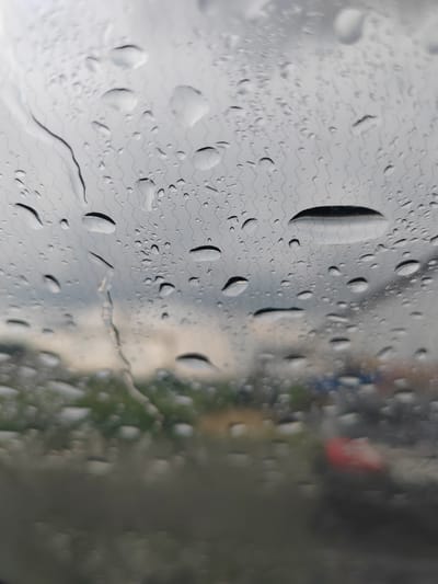 The image shows a close-up view of raindrops on a window, with a blurred outdoor scene in the background. The raindrops create a distorted and abstract view of the environment. In the background, there are indications of greenery, likely trees or bushes, and what appear to be vehicles, with one partially visible red object that could be the tail light of a car. The sky is overcast and grey. The perspective suggests the photo was taken from inside a vehicle looking outwards. The specific location in Пригородный, Russia cannot be determined from the visual information provided.