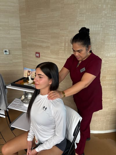 In a spa-like setting within Berry Islands, Bahamas, an adult female is performing a shoulder and neck massage on a younger female. The masseuse, identified by a "Sangeeta" name tag, wears a maroon short-sleeve uniform with her dark hair tied back. Her hands are placed on the shoulders of the seated younger female, who has long dark hair and is wearing a white long-sleeve Under Armour shirt and dark shorts. A white towel is draped over the back of the black chair the recipient is occupying. To the left, a black mobile cart holds a black tray on its upper shelf, containing smooth dark stones, a white herbal compress, yellow granular material, and two brochures, one referencing "AYURVEDA". The lower shelf holds a silver metallic bowl and several white paper sheets. The background wall is covered with light beige, horizontally-patterned tiles, featuring a white electrical outlet and a red fire alarm pull station. The flooring appears to be a light brown, smooth surface.