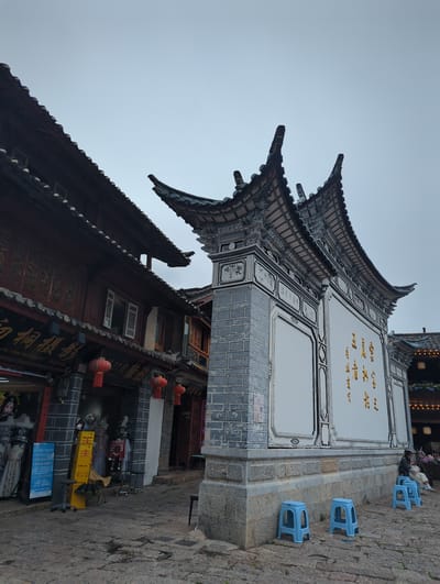 This image captures a street scene in Gucheng District, China, with a focus on traditional Chinese architecture. The foreground is dominated by the ornate, tiered roofs of buildings, characterized by their dark tiles and intricate detailing. The main building features a white facade adorned with golden Chinese characters and decorative panels, suggesting it might be a shop or a place of cultural significance.
Several bright blue plastic stools are arranged on a stone pathway in front of the building, some occupied by individuals. One person is seen sitting on a stool, appearing relaxed. The presence of these stools and the subdued lighting suggest it might be late afternoon or early evening. The sky is overcast, contributing to a muted atmosphere.
Below the main building, another structure with darker, wooden elements and a sloping roof is visible. Red lanterns hang from the buildings, adding pops of color to the scene. The cobblestone ground and the textured stone walls of the building reinforce the sense of a historic setting.
Visible text includes "CHU TONG PHOTOGRAPHY" and several lines of Chinese characters on signs and banners, some of which appear to be promotional or informational. The overall impression is of a quaint and culturally rich street in a Chinese town, likely a tourist area given the photography studio sign and the arrangement of seating.