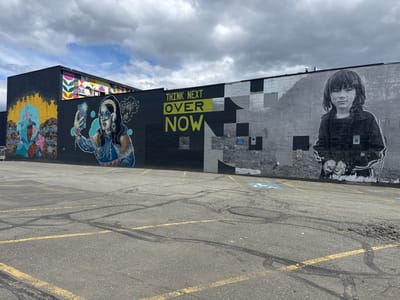 The image shows a mural on the side of a building in Anchorage, Alaska, United States. The mural depicts a variety of images, including portraits of people, abstract shapes, and text. The text reads, "Think Next Over Now." The parking lot in front of the building is cracked and has yellow lines. The sky is overcast.