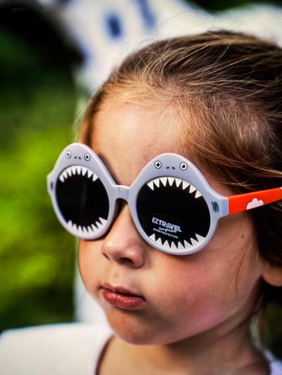 A close-up shot captures a young Caucasian child wearing novelty shark-shaped sunglasses. The sunglasses are gray with white teeth, googly eyes, and have orange earpieces with white cloud designs. The child's hair is light brown, and they are looking to the left of the frame with a neutral expression. The background is blurred, showing a mix of green foliage and what appears to be a white structure, possibly a building or furniture. The lighting suggests an outdoor setting. There is no identifiable information indicating the location in Vientiane Capital, Laos.