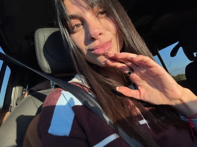 A woman with long, straight brown hair and bangs sits in a car, wearing a maroon, white, and light blue striped sweatshirt and a seatbelt. She has light skin, brown eyes, and is wearing a black ring on her left hand. Her hand is raised near her face, with her fingers slightly curved. The car's interior is visible, including a leather headrest and window. The sky is seen through the window.