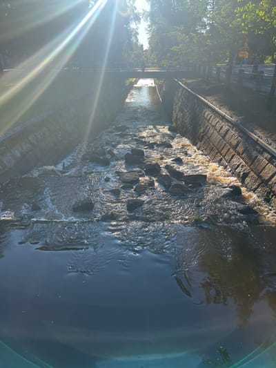 The image shows a canal with stone walls and a rocky bottom in Oulu, Finland. The water is flowing and appears somewhat murky. A bridge crosses the canal in the distance, with trees and a road visible on either side. Sunlight streams in from the upper left, creating bright lens flares and dappling the water and surrounding areas. The canal walls are constructed from rough-hewn stone blocks. There are no people or animals visible in the frame. The overall scene depicts an outdoor urban environment with natural elements.