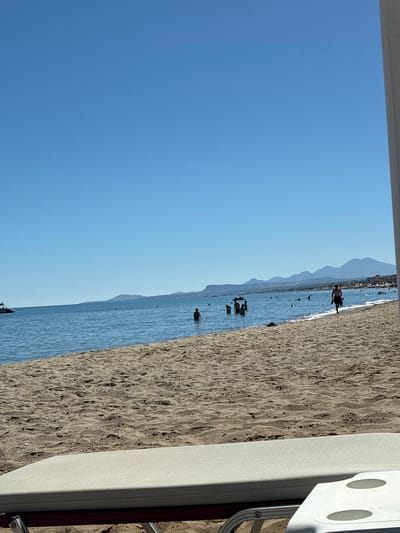 The image depicts a sunny day at a beach in Rethymno, Greece. In the foreground, a partially visible beach lounger is set on the sand. The midground features the sandy beach leading to the calm, blue sea, where several people are swimming and wading. A jet ski is visible in the far left of the water. In the background, a picturesque coastline with buildings and mountains stretches across the horizon under a clear blue sky.