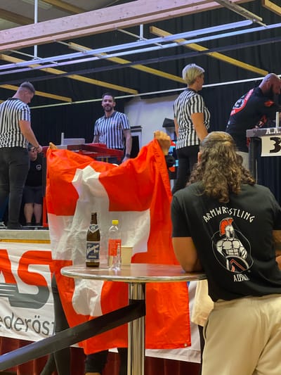 The image captures an indoor armwrestling event in Reisenberg, Austria. The perspective is from a spectator area looking towards a raised stage.
In the foreground, a male with long, curly brown hair, wearing a black t-shirt with "ARMWRESTLING AUSTRIA" and a Spartan warrior graphic, stands with his back to the viewer, leaning on a round, chrome-legged high-top table. On the table are a brown glass bottle of "Wieselburger Bier," a clear plastic bottle with a yellow cap containing a red liquid, and a white paper cup. A large red flag with a white cross, identified as the flag of Switzerland, is draped over the table and held by a hand. A black retractable stanchion belt runs below the table.
On the stage, from left to right: a male referee in a black and white striped shirt and dark pants stands next to an armwrestling table, facing away from the camera. To his right, a male referee with a beard, in a similar striped shirt, stands smiling behind an armwrestling table. Further right, a female referee with short blonde hair, also in a striped shirt, stands near another armwrestling table, facing away. A male competitor or official with a beard is partially visible on the far right, leaning over an armwrestling table marked with a white sign displaying "AA 3." Another individual in dark attire is partially visible on the left side of the stage, behind the Swiss flag.
The stage features multiple red and grey armwrestling tables. A white banner with red lettering, partially reading "AS" and "ederöste," is visible along the base of the stage. The background consists of dark curtains behind the stage. The ceiling area reveals exposed wooden beams, metal structural elements, and fluorescent lighting fixtures.