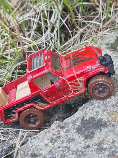 A red toy truck with black accents and mud splatters sits on a rough, grey concrete surface. The truck is oriented diagonally across the frame, with its front facing towards the lower right corner. It is partially surrounded by dry, light-colored grass and weeds. The concrete appears to be outdoors, possibly in an urban or semi-urban environment. The scene is lit by natural daylight, casting subtle shadows. The overall condition of the toy truck suggests it has been used outdoors, experiencing some wear and tear. The location of Žygaičiai, Lithuania, is provided as context for this outdoor scene. There are no people visible in the image.