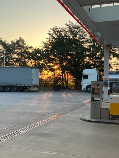 The image depicts a gas station located in Gorni Dabnik, Bulgaria, during a sunset. A large, white semi-truck is parked on the asphalt lot, its trailer facing left. To the right, another white semi-truck is positioned near a gas pump, with a yellow and white fuel dispenser visible. In the background, a dense line of trees with lush green foliage creates a natural barrier, with the setting sun casting a warm, golden glow and long shadows across the scene. The sky above is a gradient of soft yellow and pale blue, indicating the approaching evening. The ground in the foreground is asphalt, with a drainage grate running diagonally across it. A grey trash receptacle is situated near the gas pump. The overall atmosphere is serene and quiet, suggestive of a brief pause in transit.