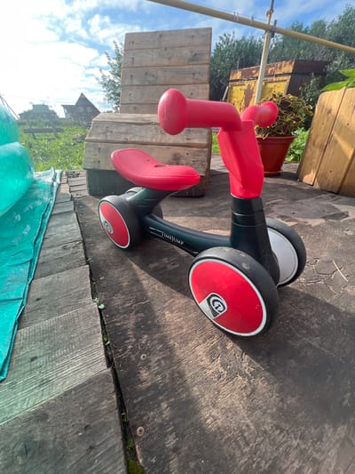 The image shows a red and black children's ride-on toy in a yard with wooden structures and greenery. The toy has a red seat and red handlebars with black and red wheels. It is positioned on a wooden surface, possibly a deck or pathway, which is weathered and has visible grain. To the left of the toy, a large teal tarp is visible, partially covering the ground. In the background, there are wooden planks stacked vertically, a potted plant with red and green foliage, and a rusty metal structure. Further in the distance, houses and trees are visible under a bright blue sky with scattered clouds. The scene suggests an outdoor setting, possibly a backyard in the village of Нестюково, Russia, given the presence of residential structures in the background and the overall rural or semi-rural aesthetic. There are no people or animals present in the image.