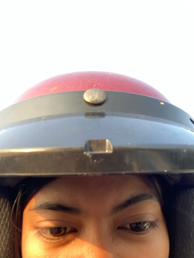 This is a close-up, vertically oriented photograph of a person's eye and part of a red and black helmet. The person's eye is dark brown with visible eyelashes and eyebrows. The skin tone appears to be light to medium. The helmet has a rounded red section and a darker, possibly black or grey, segmented section. A metallic button or rivet is visible on the red part of the helmet. The background is a bright, blown-out white, suggesting an overexposed sky or a very bright, uniform light source. The lighting on the person's face suggests it is daytime. The image quality is somewhat grainy and appears to have been taken at a close proximity. There is no visible text in the image. The composition is tightly cropped, focusing on the eye and the helmet.