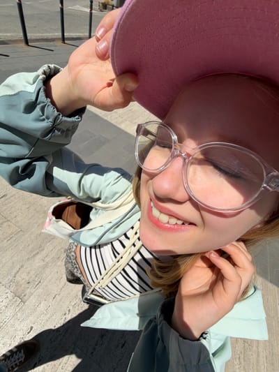 A photograph, taken from above, shows a fair-skinned woman smiling with her eyes partially closed. She is wearing clear-framed glasses and a pink hat. She has fair hair. She is wearing a striped shirt under a jacket. The jacket is light blue with grey sleeves. She is holding the edge of the pink hat. The background shows a beige-colored cobblestone surface. In the bottom right corner, a single foot is visible in a brown shoe. The location appears to be San Gimignano, Italy, given the stone paving material. The photograph appears to have been taken in daylight.