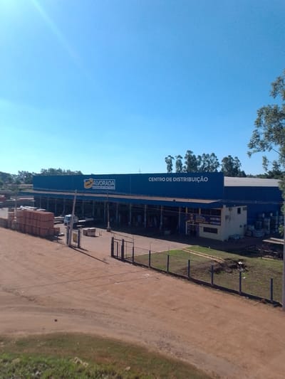 This is an exterior, high-angle, wide shot of a large, blue commercial building in Campo Grande, Brazil. The building appears to be a distribution center for construction materials, as indicated by the signage that reads "ALVORADA MATERIAIS DE CONSTRUÇÃO" and "CENTRO DE DISTRIBUIÇÃO". The building has a long, flat roofline and is supported by multiple blue pillars along its facade. Stacks of reddish-brown bricks and other building materials are visible outside the structure, particularly on the left side of the image. A white truck with a flatbed trailer is parked in front of the building. The surrounding area is a mix of dirt, grass, and a few trees. The sky is clear and blue. A chain-link fence with barbed wire runs along the perimeter of the property, and a metal gate is visible on the right side of the image. The scene suggests a sunny day at a commercial facility.