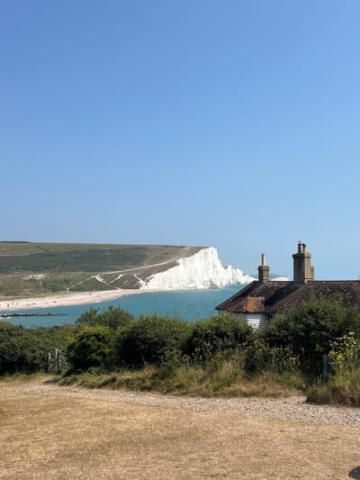 The image depicts a scenic coastal view of the Seven Sisters cliffs in Seaford, United Kingdom. In the foreground, dry, sun-bleached grass covers the ground, with a gravel path leading towards a wooden gate and dense green shrubbery. Partially visible behind the foliage is a white building with a brown tiled roof and two chimneys. The middle ground is dominated by the turquoise sea, which meets a pebble beach dotted with a few small figures. Beyond the beach, the iconic white chalk cliffs of the Seven Sisters stretch into the distance, meeting a clear blue sky. The overall impression is one of a bright, sunny day in a coastal landscape.