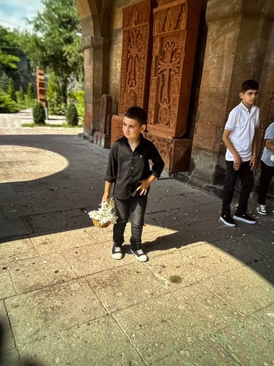 In Artik, Armenia, a young boy stands on a stone-paved walkway outside a building adorned with carved stone reliefs. He is wearing a black button-down shirt and black pants, and holds a small woven basket filled with white flowers. To his right, another boy in a white polo shirt and black pants stands looking away. The background shows architectural elements and greenery, suggesting a historical or religious site. The sun casts strong shadows on the ground.