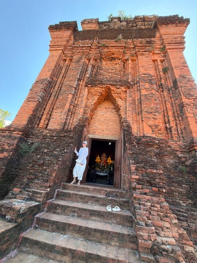 A woman in a white outfit and white cap stands on a brick staircase in front of a large, ancient brick tower in Tuy Hoa Ward, Vietnam. The tower has a wide, arched entrance where an altar with yellow flowers and statues is visible. Two pairs of sandals are placed on the steps. The sky is clear and blue.