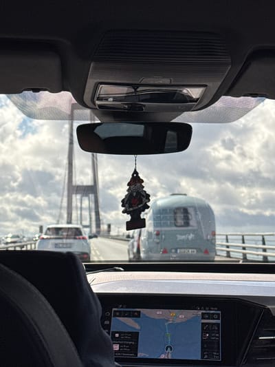 The image shows a view from inside a car, looking forward through the windshield. A car air freshener hangs from the rearview mirror. In front of the car, traffic is moving across a large bridge. A light blue camper is in the right lane, and a white car is in the left lane. The bridge appears to be the Great Belt Bridge, which connects the Danish islands of Zealand and Funen. The Great Belt Bridge is located in Korsør, Denmark. The sky is overcast with scattered clouds. The car's dashboard display is visible in the foreground, showing navigation information.