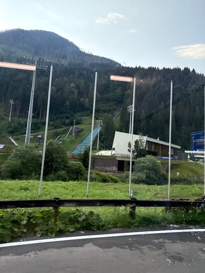 The image captures a ski jumping complex nestled within a mountainous, forested region. The ski jumps, with their steep inclines and green landing surfaces, are visible on the hillside. A building, likely a lodge or administrative center, stands at the base of one of the jumps. Several tall poles with lights are interspersed throughout the scene, suggesting the facility can be used at night. The environment is predominantly green, with lush grass covering the slopes and dense trees forming a backdrop. The sky is partly cloudy, with a clear blue expanse visible in the upper portion of the frame. The image appears to be taken from a moving vehicle, indicated by the blurred foreground and the presence of a guardrail. The ski jumping complex is located in Predazzo, Italy, a town known for its winter sports facilities.