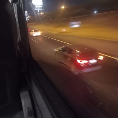 The photo appears to be taken from inside a vehicle, possibly a bus or large van, at night. Out the front window, blurred lights and signs suggest a highway or city street scene. Streetlights are visible on the left, and the headlights and tail lights of other vehicles are visible in the distance. A red object, possibly a plastic cup or bottle, is adhered to the ceiling of the vehicle. The interior of the vehicle is dimly lit.
