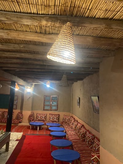 This is an interior scene of a traditional Moroccan tent or riad in Agafay, Morocco. The ceiling is constructed from exposed wooden beams and bamboo poles, with a large, woven conical lamp hanging from the center. The walls are made of natural clay or mud plaster, giving the space a rustic feel. Along the walls are built-in benches covered with red and white patterned cushions, creating a comfortable seating area. In front of the benches are low, round blue tables. Several other woven lamps are visible, as well as framed artwork on the walls. Red carpets cover the floor, adding warmth and color to the space. The overall ambiance is one of a cozy, authentic Moroccan dwelling, likely a restaurant or guesthouse.