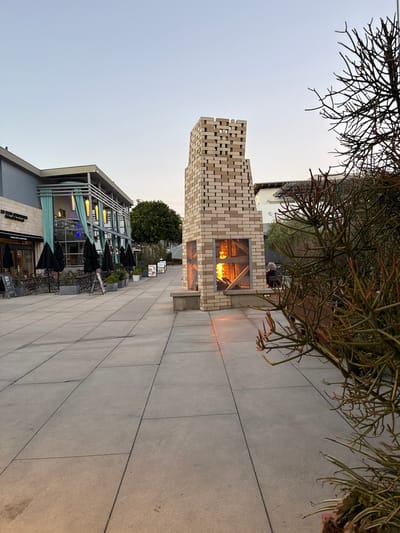 An outdoor dining area in Manhattan Beach, California, features a tall, stacked-brick fireplace with a lit fire. The fireplace is situated on a concrete patio, with a single person seated on a bench facing away from the camera. To the left, a building with a modern design has outdoor seating arranged with black umbrellas. In the foreground, a spiky, leafless plant obscures part of the view. The sky is clear and light blue, indicating it is either early morning or late evening.