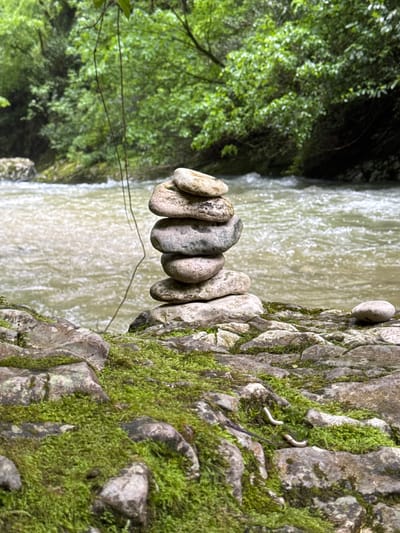 The image depicts a small stack of balanced stones on a moss-covered rock face next to a waterfall in Tsabal, Abkhazia. The stones are smooth and various shades of grey. Lush green foliage is visible on the left of the image, contrasting with the turbulent, light brown water of the waterfall in the background. The moss on the rock face is dense and vibrant green. A twig spans the gap between the stone stack and the foliage.