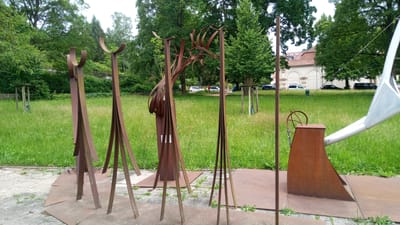 The image depicts an outdoor sculpture installation on a paved area next to a grassy lawn. In the foreground, there are several abstract metal sculptures, primarily made of rusted metal. These sculptures are tall and slender, with geometric and flowing shapes. One sculpture on the far left has two curved elements at the top, resembling stylized horns or ears. To its right, another sculpture has a more complex arrangement of thin, curved metal pieces extending upwards. Further to the right, there is a sculpture that resembles a watering can, made of rusted metal, with a circular element on top and a silver-colored spout extending towards the right edge of the frame. Behind the sculptures, a wide expanse of green grass is visible, interspersed with small yellow wildflowers. Beyond the lawn, a paved area with parked cars can be seen. In the background, there is a building with a red-tiled roof and white arched facades. Trees with lush green foliage are scattered throughout the scene. The sky is overcast and light. The location is the Lutherhaus in Eisenach, Germany.