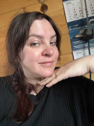 A woman with brown hair and fair skin poses for a selfie. She is wearing a black top. Her right hand is resting on her face, showing a ring on her finger. In the background, there is a calendar hanging on a wooden wall. The calendar shows the year 2026 and lists "Почтовый адр г. Пермь ул. Кра" and "Юридический адр г. Пермь, ул. Маш". This information indicates a connection to the city of Perm, Russia, rather than Bol'shakino. A black power cord and adapter are visible above the calendar.