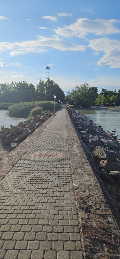 The image depicts a paved walkway, likely a pier or promenade, leading towards a body of water. The walkway is constructed from grey and reddish-brown interlocking bricks, bordered on one side by large, rough-hewn rocks and on the other by water. Along the walkway, several ornate lampposts are visible, spaced at intervals. In the background, lush green trees and foliage line the shore. A cell tower with a cage-like structure at its top stands prominently against a bright blue sky with scattered white clouds. A few people are seen walking along the path and in the distant background, suggesting a public recreational area. On the right side, the water's edge is also lined with rocks, and a couple of ducks are swimming in the water. The overall scene appears to be a scenic view of a lakeside or riverfront area, possibly in Balatongyörök, Hungary, known for its attractive waterfronts.
