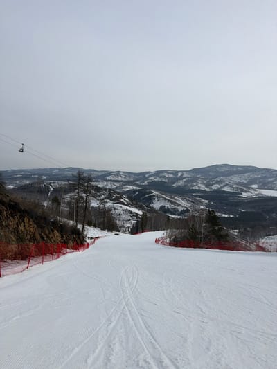 The image displays a snow-covered ski slope within a mountainous landscape at a ski resort in Novoabzakovo, Russia. The sky is uniformly overcast and light grey. A wide, groomed ski run dominates the foreground, showing distinct parallel tracks from a snow groomer on the left half and multiple ski/snowboard tracks further right. Red safety netting delineates both sides of the ski path. To the left, a rocky, snow-dusted embankment rises, featuring several bare deciduous trees. To the right, a mix of snow-dusted evergreen and bare deciduous trees lines the slope. In the upper left quadrant, a single, empty ski lift chair is suspended by cables, moving across the frame. The background consists of undulating snow-covered mountains and hills, partially forested with coniferous and deciduous trees. No individuals are present in the scene.