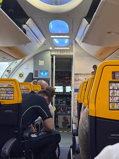 The image shows the interior of a Ryanair airplane, with a view towards the cockpit. Passengers are seated in the yellow seats, and overhead compartments are visible. Signage includes "EXIT," "LIFE VEST IN PANEL ABOVE YOUR HEAD," "FASTEN SEAT" and "RYANAIR" are visible. The image suggests the aircraft is likely in flight or preparing for take-off or landing. There is no information to determine the location is in the city of Granadilla de Abona, Spain.