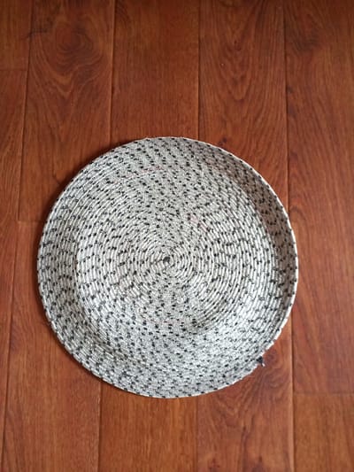 A circular, woven mat with a speckled grey and black pattern rests on a polished wooden floor. The mat is positioned slightly off-center in the frame, with its bottom edge visible. The floorboards are a warm brown color and exhibit a wood grain pattern. The scene appears to be indoors, likely in a domestic setting. There is no information in the image to suggest a location in Vilyuysk, Russia.