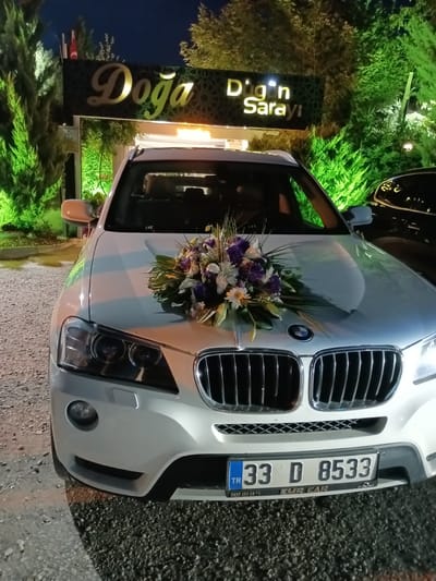 The image shows the front of a white BMW X3 parked at night. The car has a floral arrangement on its hood. Behind the car, a sign reads "Doğa Düğün Sarayı" which translates to "Nature Wedding Palace". The location is the city of Kırıkkale, Turkey. The background consists of trees illuminated with green lights. The ground is covered with gravel. The license plate of the car is visible and reads "33 D 8533".